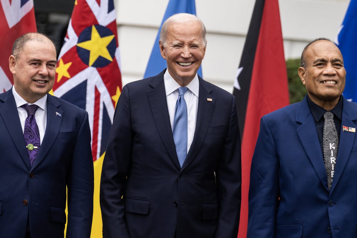 Three persons in suits smiling in front of flags